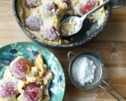 A portion of roasted plum clafoutis with the remainder in the pan, dusted with icing sugar
