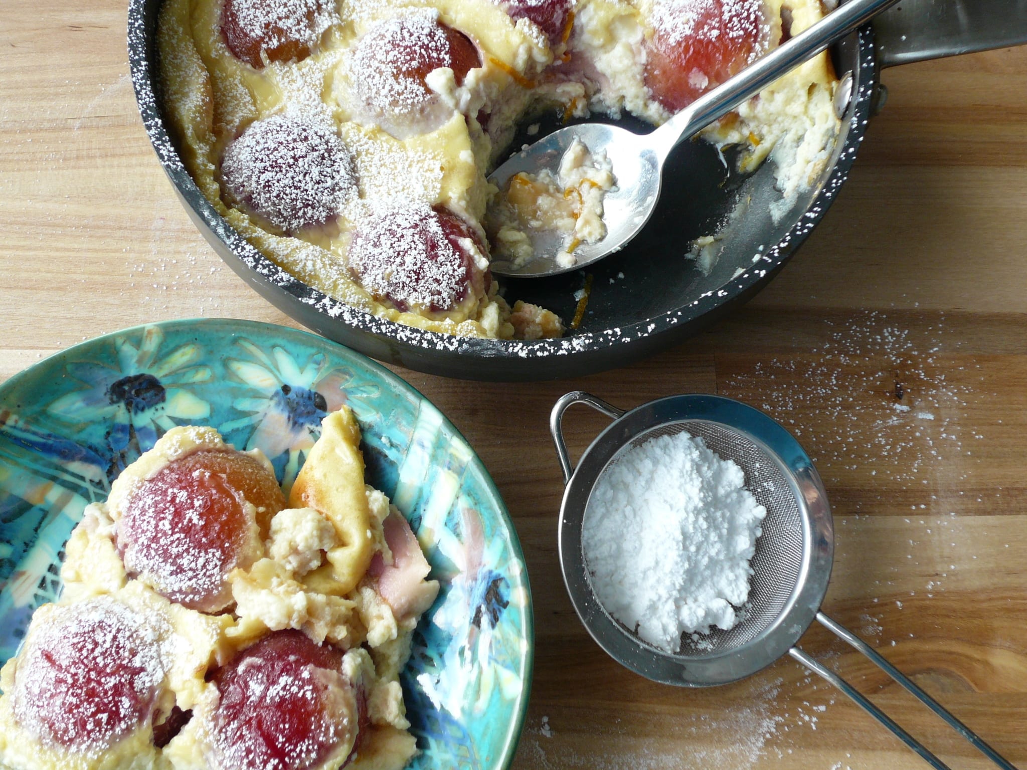 A portion of roasted plum clafoutis with the remainder in the pan, dusted with icing sugar